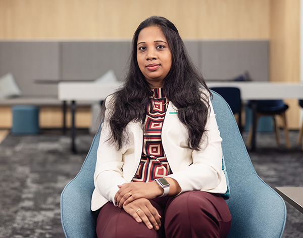 A woman with long dark hair, wearing a white blazer and patterned top, sits in a modern office chair with her hands folded in her lap, looking at the camera. The background shows a contemporary office setting.
