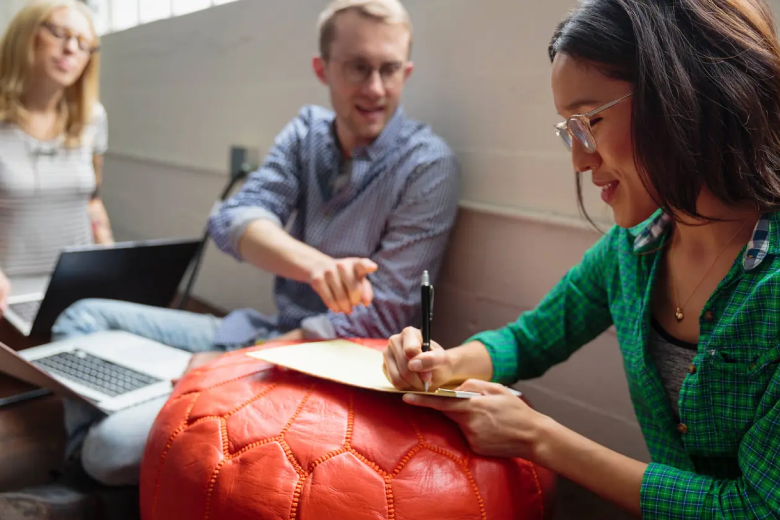 Three people sit together in a casual setting. One woman writes on a clipboard while smiling, a man gestures and talks to her, and another woman works on a laptop in the background.