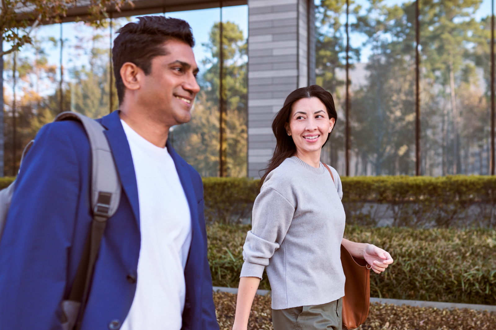 Two people walk outdoors near a modern building with large glass windows. The man, carrying a backpack, wears a blue blazer and white shirt. The woman, holding a bag, smiles at him. Trees and shrubs are in the background.
