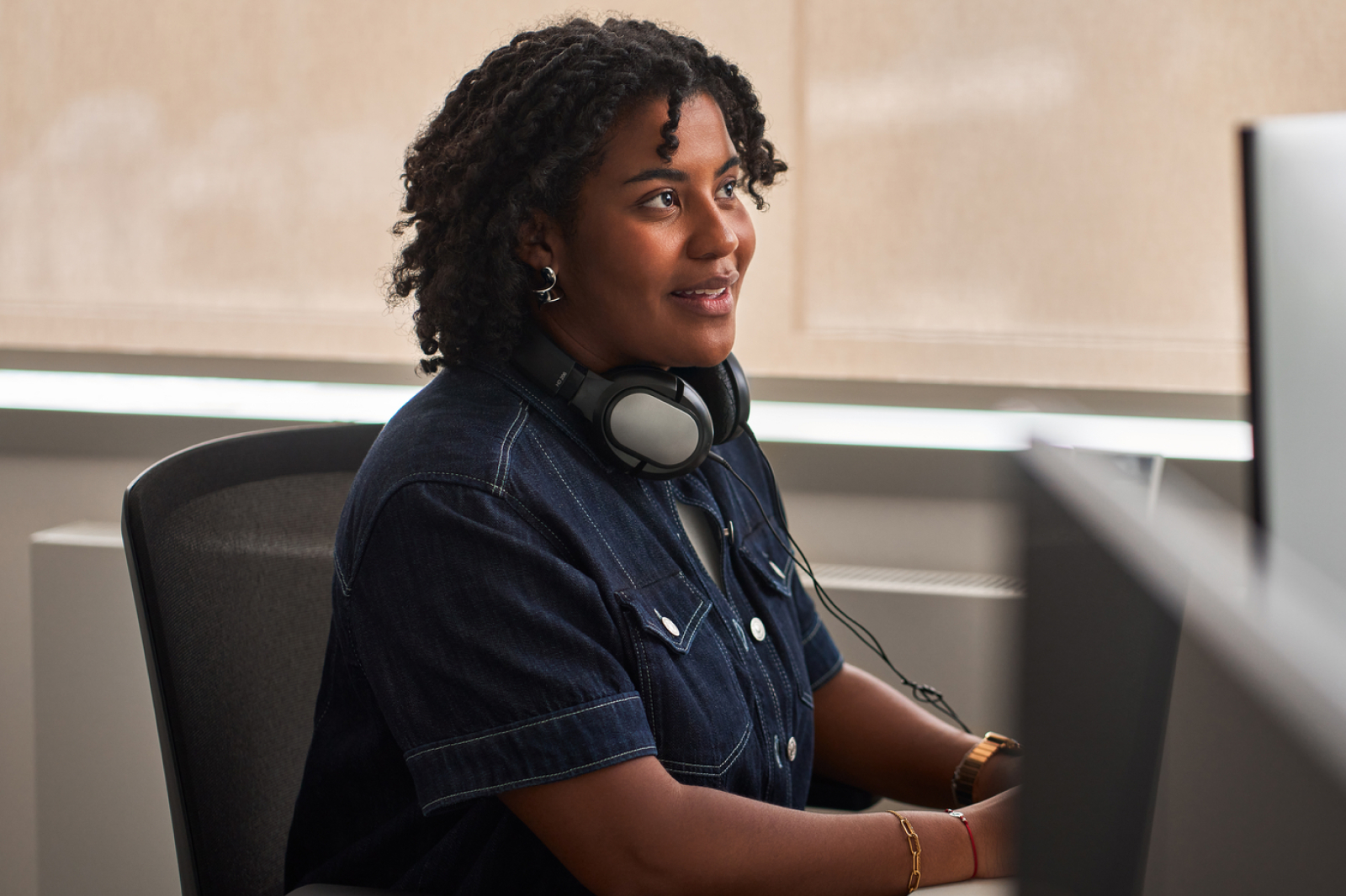 A woman with curly hair, wearing a dark denim shirt and headphones around her neck, sits at a desk and looks at a computer screen with a focused expression.