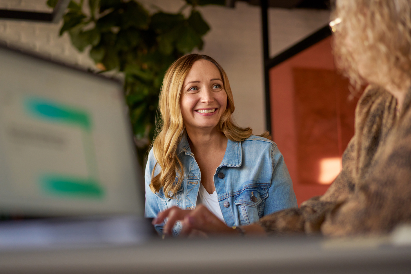 A woman with long blonde hair in a denim jacket smiles while sitting at a table. Another person sits across from her, partially visible, and a laptop is open in the foreground.