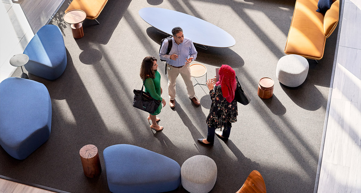 Three people stand talking in a modern lounge area with contemporary furniture, including blue and orange seats and round stools, as sunlight streams in through large windows.