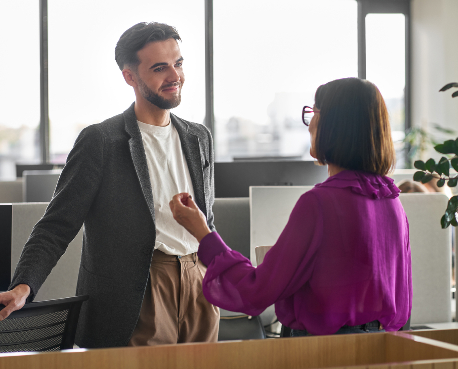 Two people stand and have a conversation in a modern office. One person wears a dark blazer and white shirt, while the other wears a purple blouse and glasses. They are smiling and appear to be engaged in discussion.