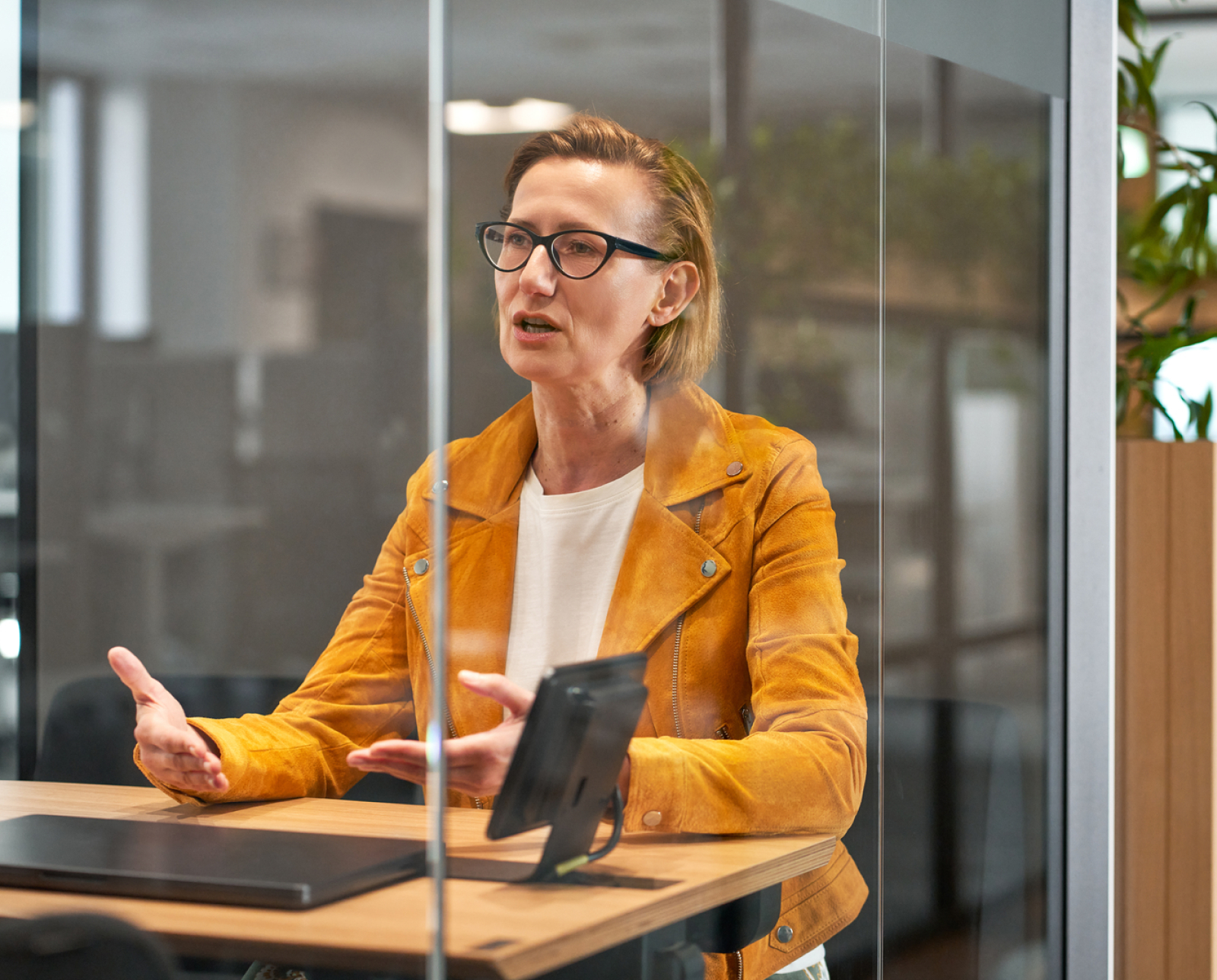A woman wearing glasses and a mustard yellow jacket sits at a desk in a modern office, gesturing with her hands while speaking. A tablet and notebook are on the desk in front of her.