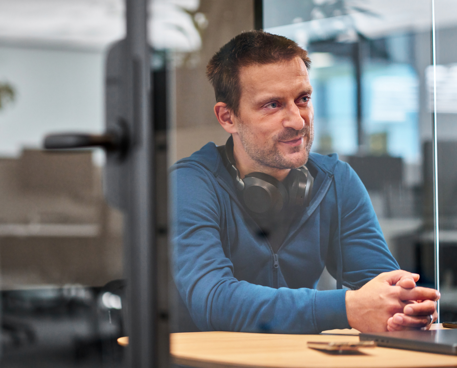A man with short brown hair, wearing a blue zip-up hoodie and headphones around his neck, sits at a table in a modern office, looking to the side and smiling.