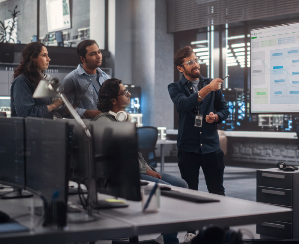 A group of four people in an office are gathered around a large screen displaying a flowchart. One person is pointing at the screen while the others listen attentively. Computer monitors and technical equipment are visible.