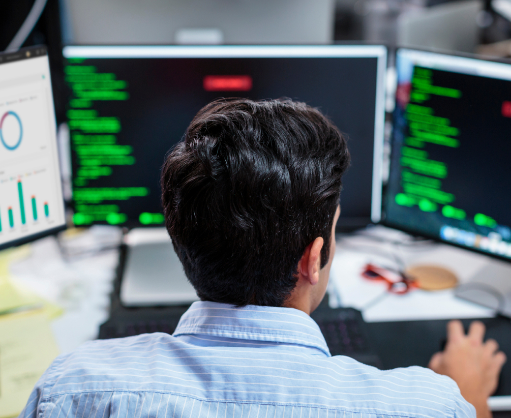 A person with dark hair sits at a desk, facing three computer monitors displaying code, graphs, and data, suggesting they are working on programming or data analysis in an office environment.