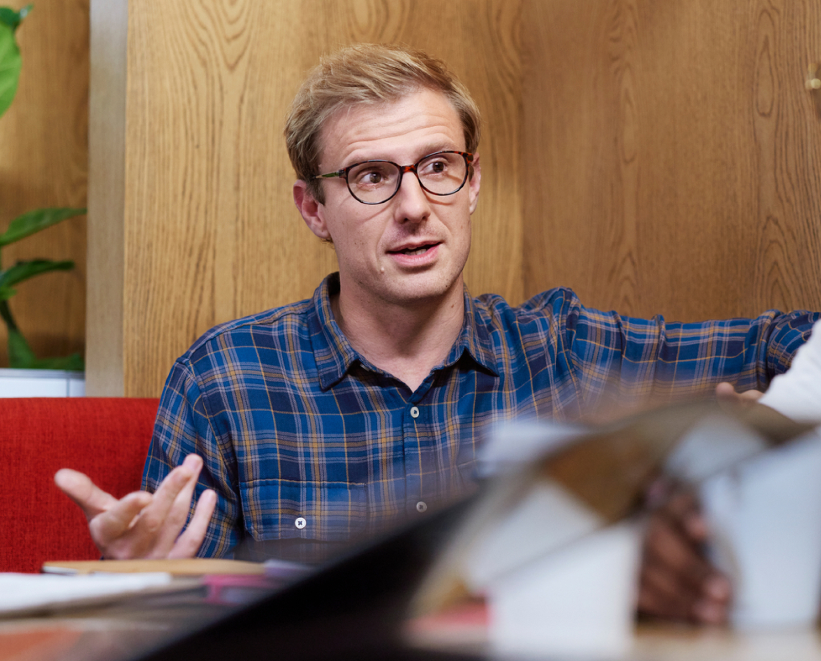 A man with blond hair and glasses, wearing a blue plaid shirt, sits indoors and gestures with his hands while speaking. There are books and papers in the foreground.
