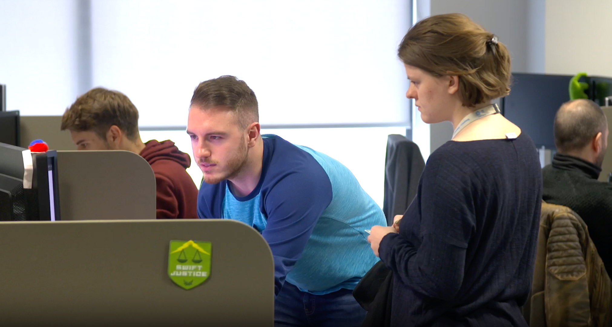 Three people work in a modern office. One man leans over his desk, focusing on his computer, while a woman stands beside him watching. Another man sits at a nearby desk, also using a computer.