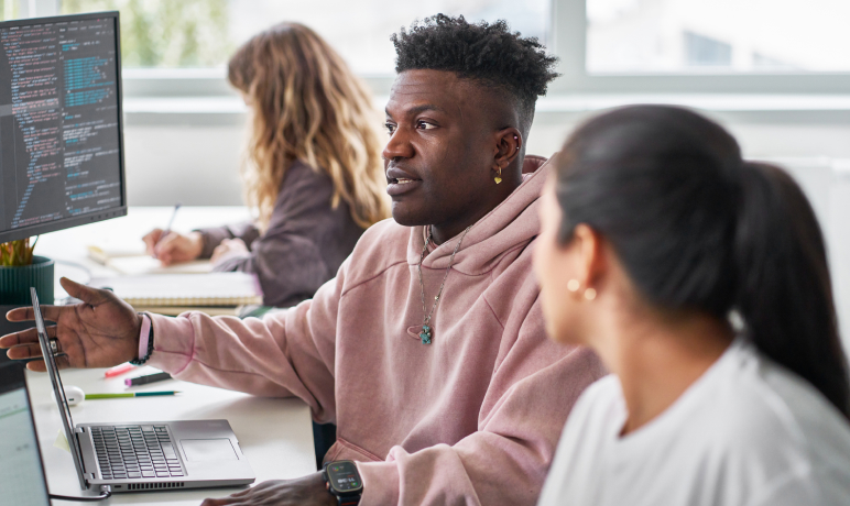 A man in a pink hoodie talks to a coworker at a desk with laptops and computer monitors displaying code, while another person writes in a notebook in the background.