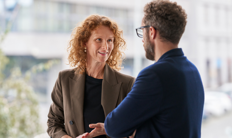 A woman with curly red hair and a brown blazer smiles while talking to a man with glasses and a dark blue jacket, indoors near large windows.