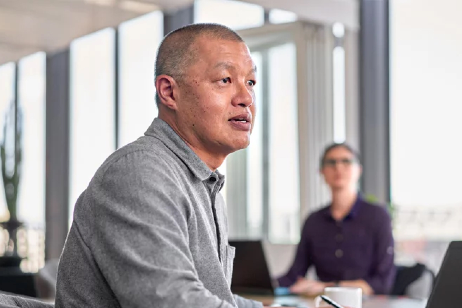 A man in a gray shirt sits at a table in a bright office, speaking and looking to the side. In the background, another person with glasses is seated, slightly out of focus, with large windows letting in natural light.