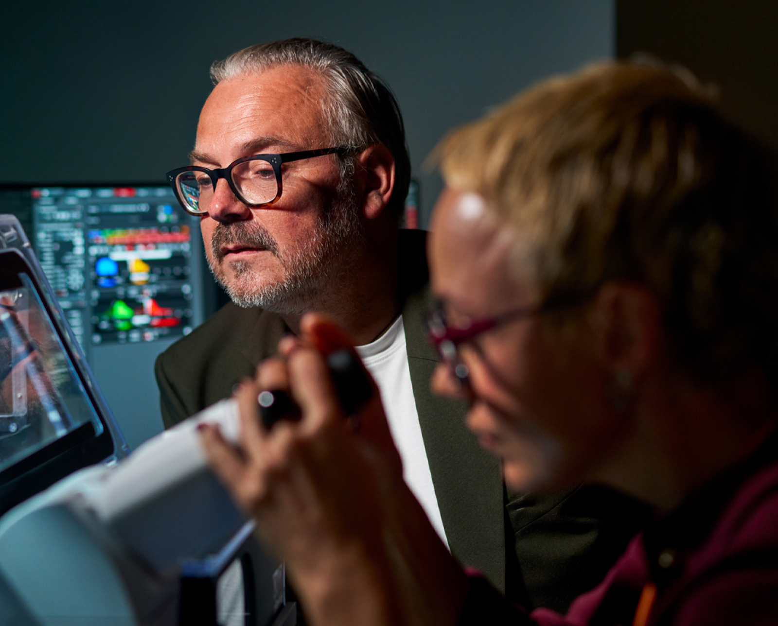 Two scientists in a lab, one observing data on a computer screen and the other using a laboratory microscope, both wearing glasses and focused on their work.