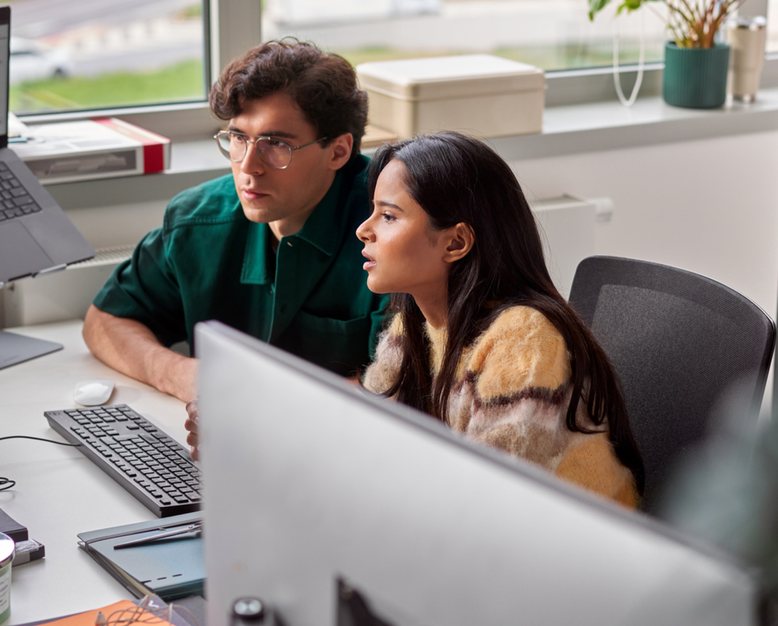 Two people sit closely together at a desk, looking attentively at a computer screen. Office supplies and a window with plants are visible in the background, creating a modern workspace atmosphere.
