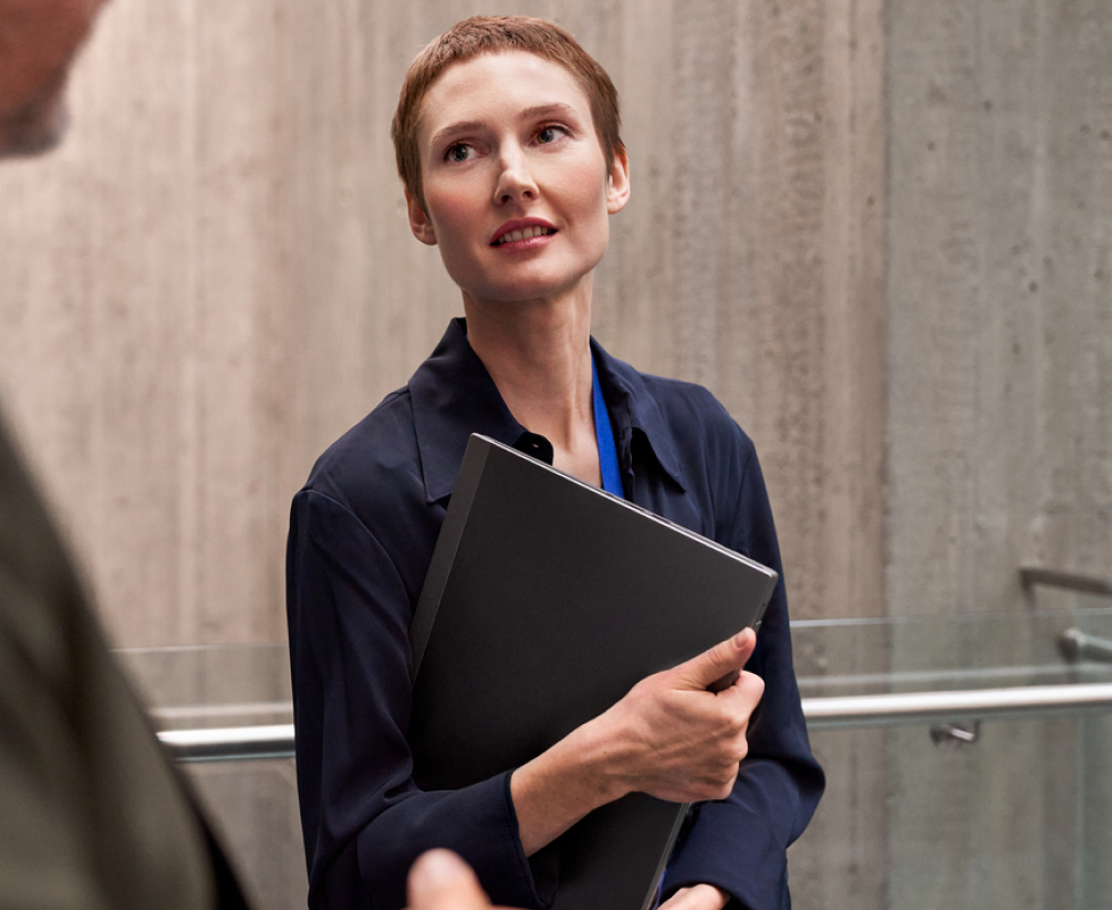 A person with short hair, wearing a dark shirt, holds a black folder while standing indoors near a concrete wall, looking up and smiling slightly at someone out of frame.