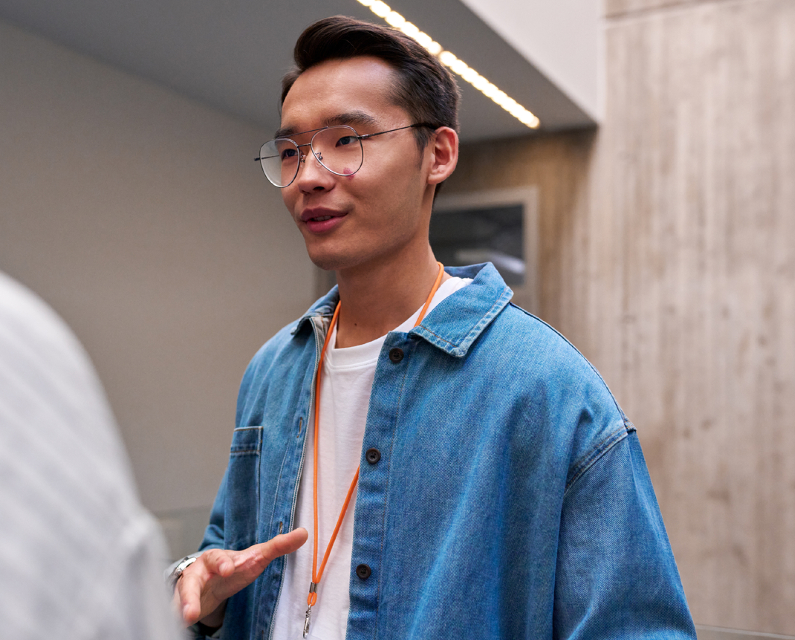 A person wearing glasses, a blue denim jacket, and an orange lanyard speaks to someone indoors, gesturing with one hand. The background includes a light-colored wall and part of a ceiling.