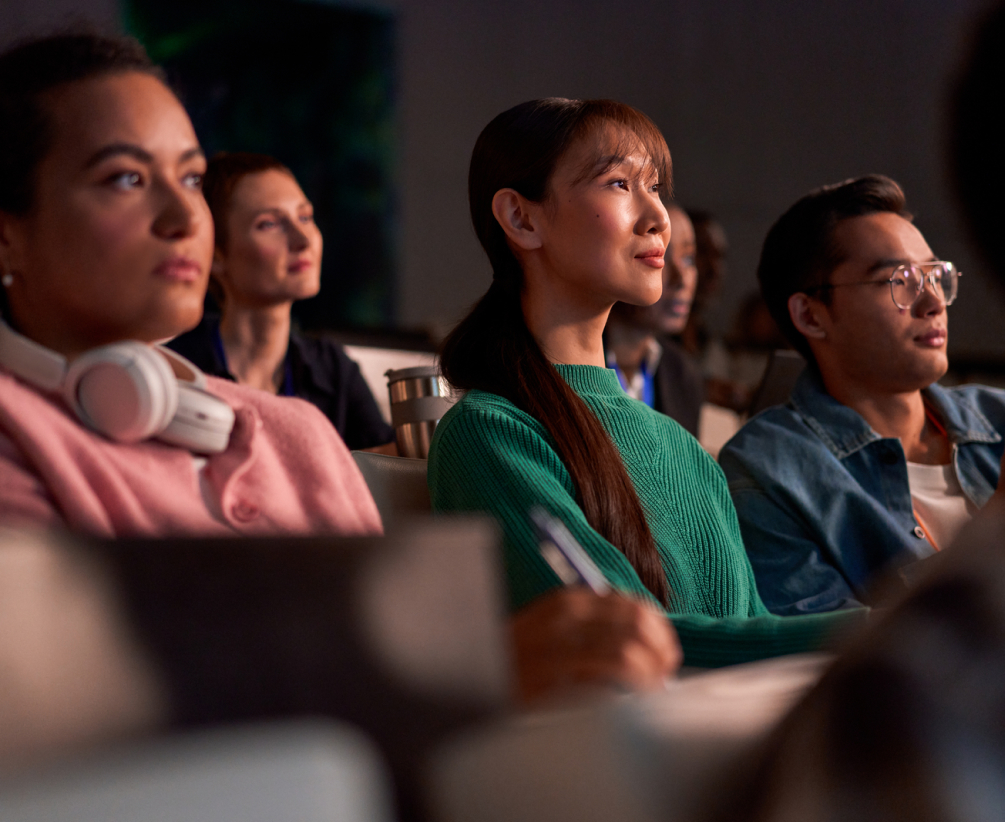 A group of young adults sit closely together in a classroom, attentively watching and listening to something ahead of them. One person takes notes while others appear engaged and focused.