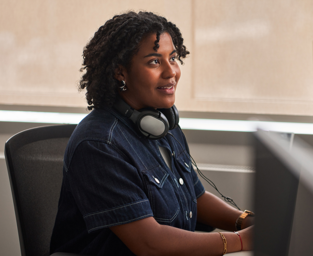 A woman with curly hair, wearing a dark denim shirt and headphones around her neck, sits at a desk and looks attentively at a computer screen.