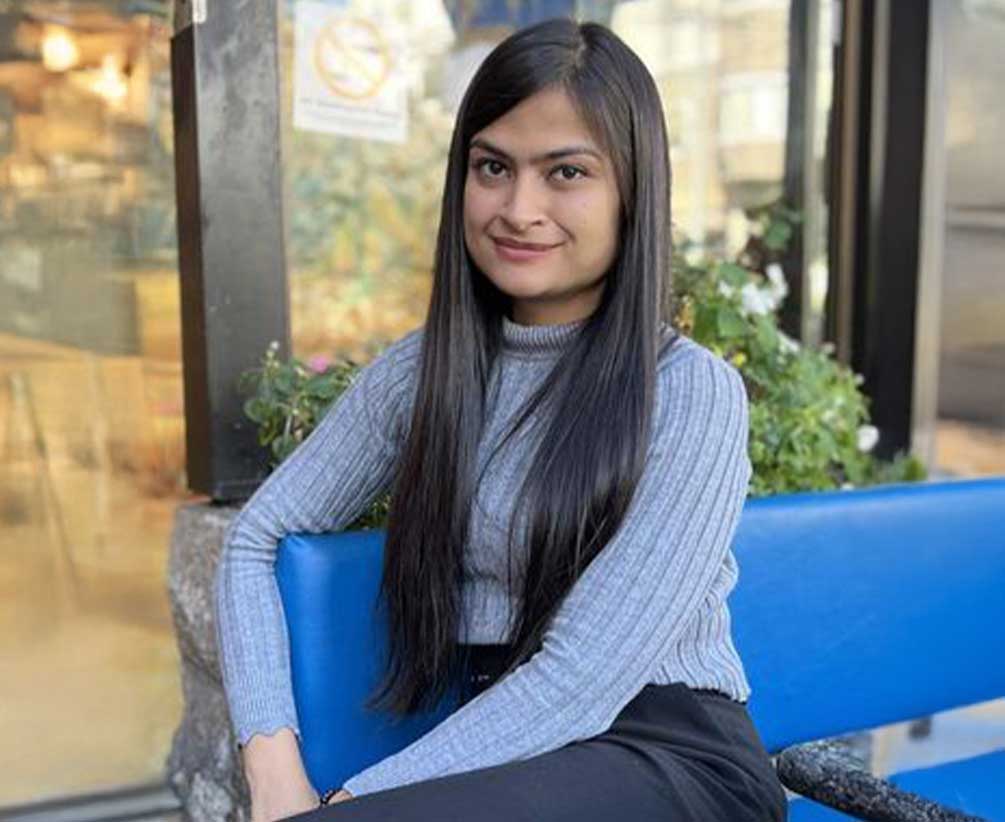 A woman with long, straight black hair sits on a blue bench outdoors, wearing a gray ribbed turtleneck sweater and black pants. She is smiling slightly, with greenery and a glass window in the background.