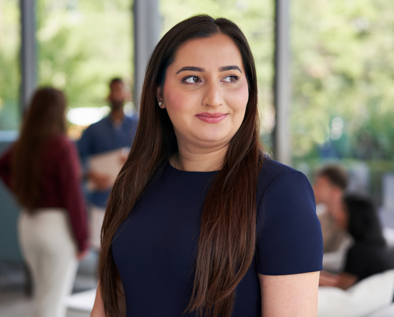 A woman with long brown hair, wearing a navy blue top, stands indoors and smiles slightly while looking to the side. Blurred people and greenery can be seen in the background.