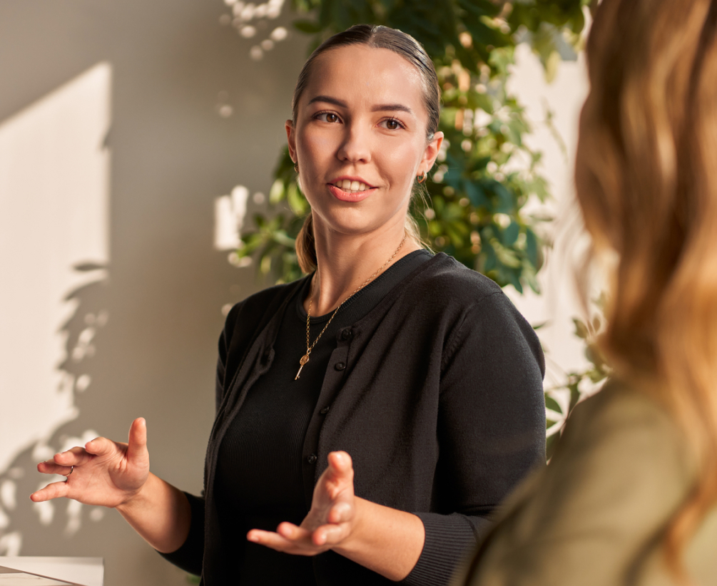 A woman with dark blonde hair tied back is speaking and gesturing with her hands indoors, facing another person whose back is to the camera. There is greenery and sunlight in the background.