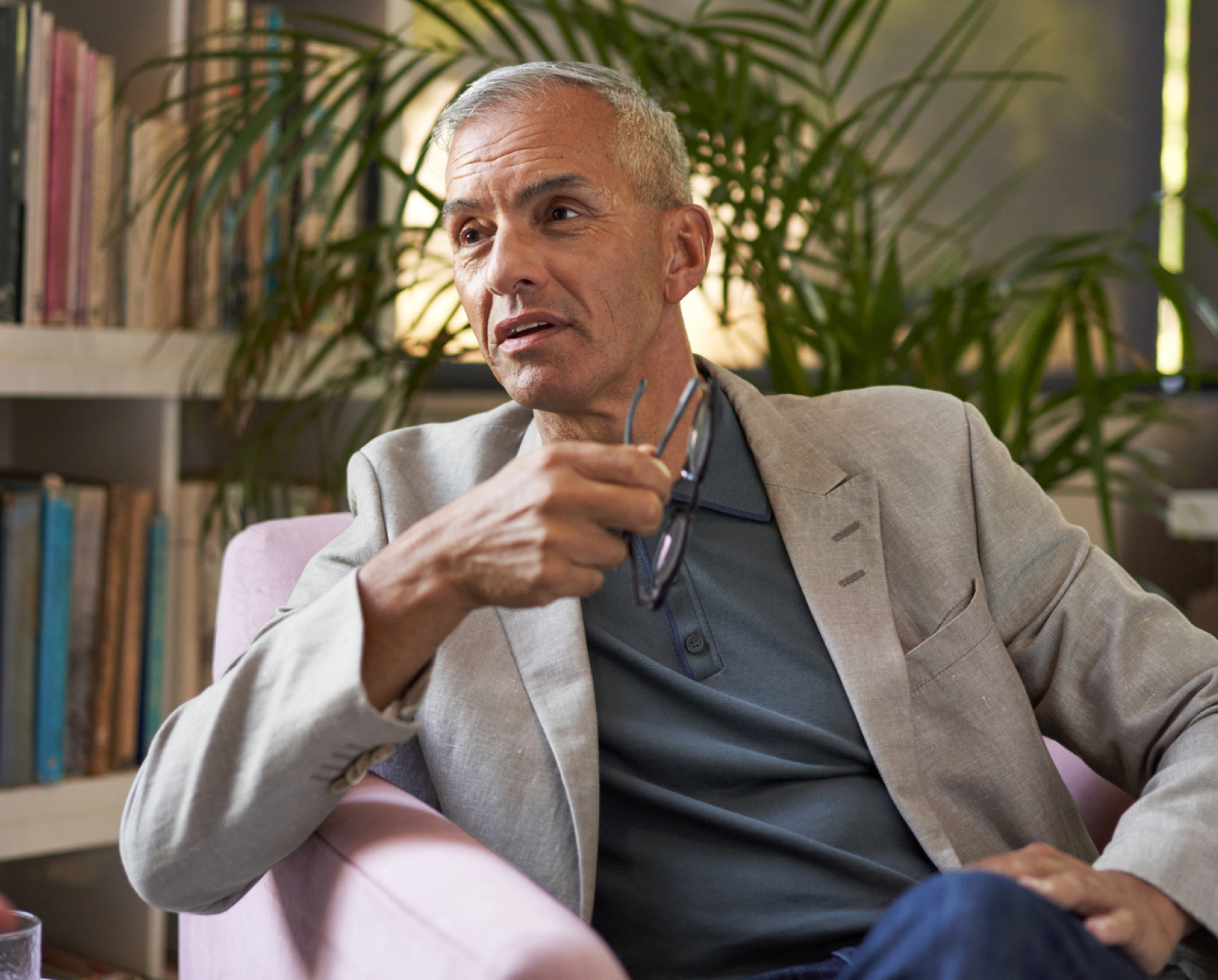 A middle-aged man with short gray hair sits in a pink chair, holding eyeglasses in one hand, wearing a gray blazer and blue polo shirt, with bookshelves and plants in the background.