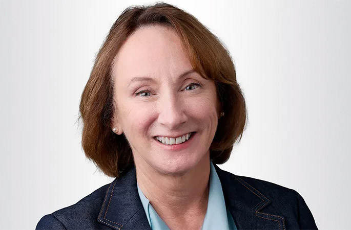 A woman with shoulder-length brown hair smiles at the camera. She is wearing a dark blazer over a light blue blouse and is posed in front of a plain, light background.