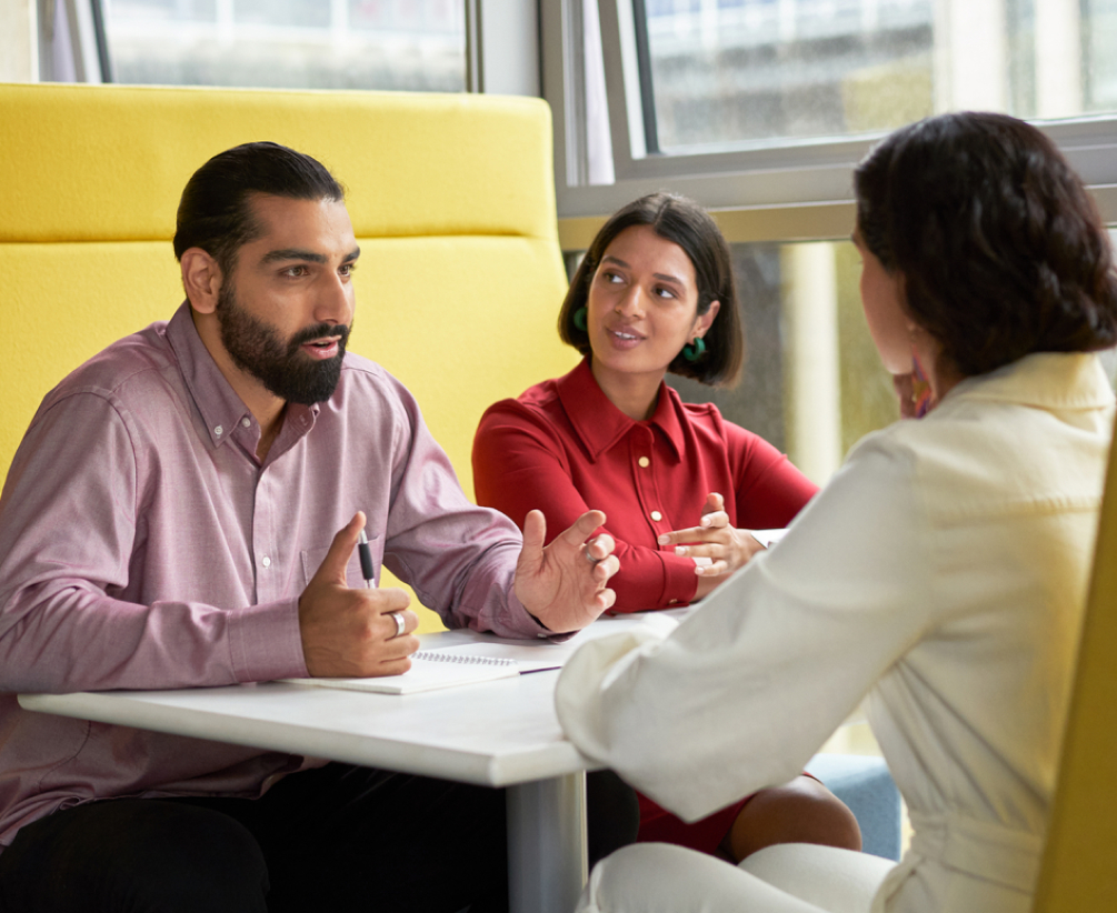Three people sit at a table in a bright office space, engaged in discussion. One man gestures while speaking, and two women listen attentively. A notebook and pen are on the table.