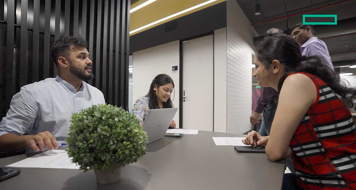 Four people are gathered around a table in an office setting, engaged in discussion. One person is using a laptop, and papers are spread on the table. A green potted plant is in the foreground. Two more people stand in the background.
