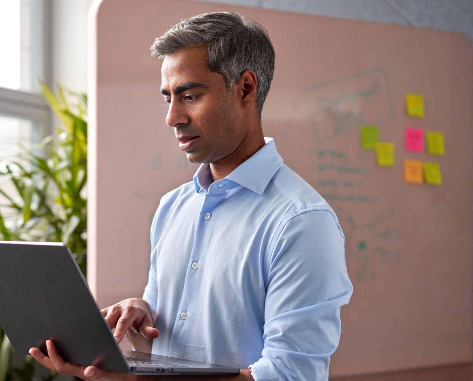 A man in a light blue shirt types on a laptop in an office. Behind him is a whiteboard with diagrams and colorful sticky notes, and a green plant is near a window.