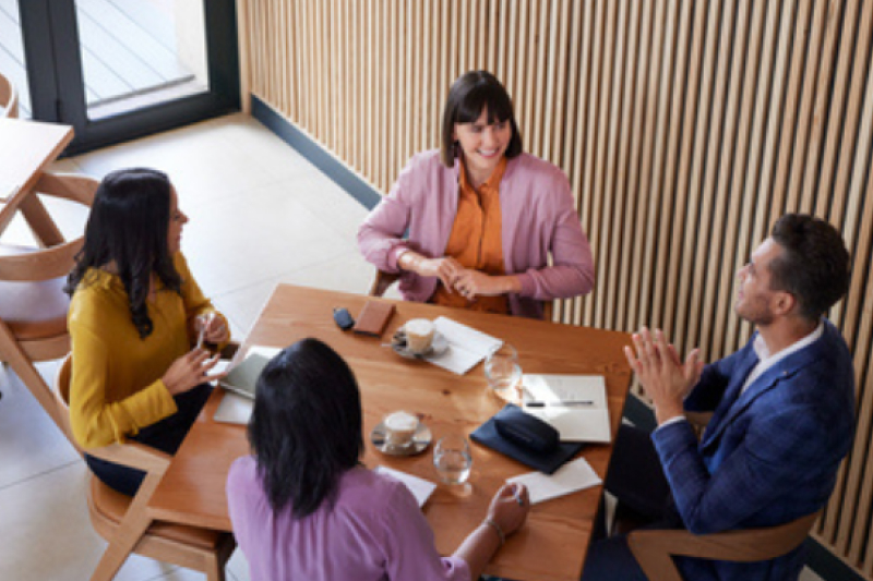 Four people sit around a wooden table in a modern cafe, engaged in conversation. They have notebooks, drinks, and phones on the table, suggesting a casual meeting or discussion.