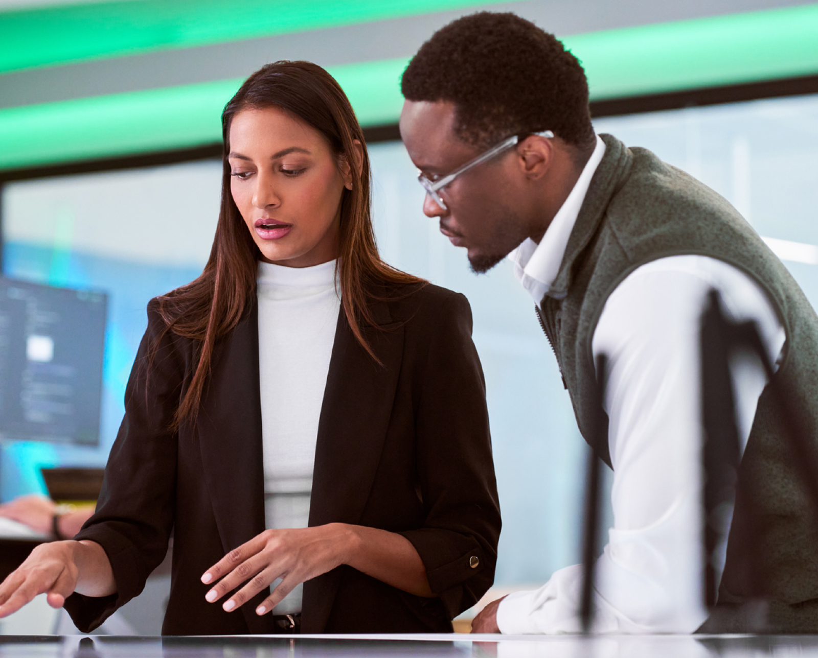 Two professionals, a woman and a man, stand together in an office, looking at documents or a digital screen on a desk and discussing work. The background features modern lighting and computer monitors.