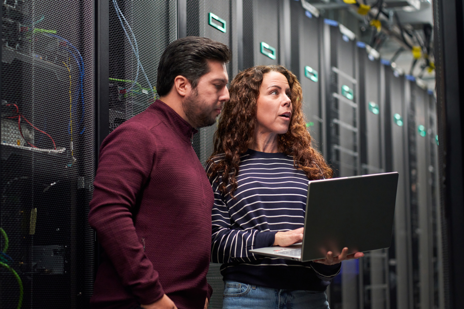 Two people stand in a server room; the woman holds an open laptop and speaks while the man looks at the screen. They appear to be discussing something technical among tall server racks.