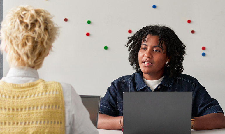 Two people sit across from each other at a table with laptops. One person is facing the camera, listening attentively, while the other has their back to the camera. A whiteboard with colorful magnets is in the background.
