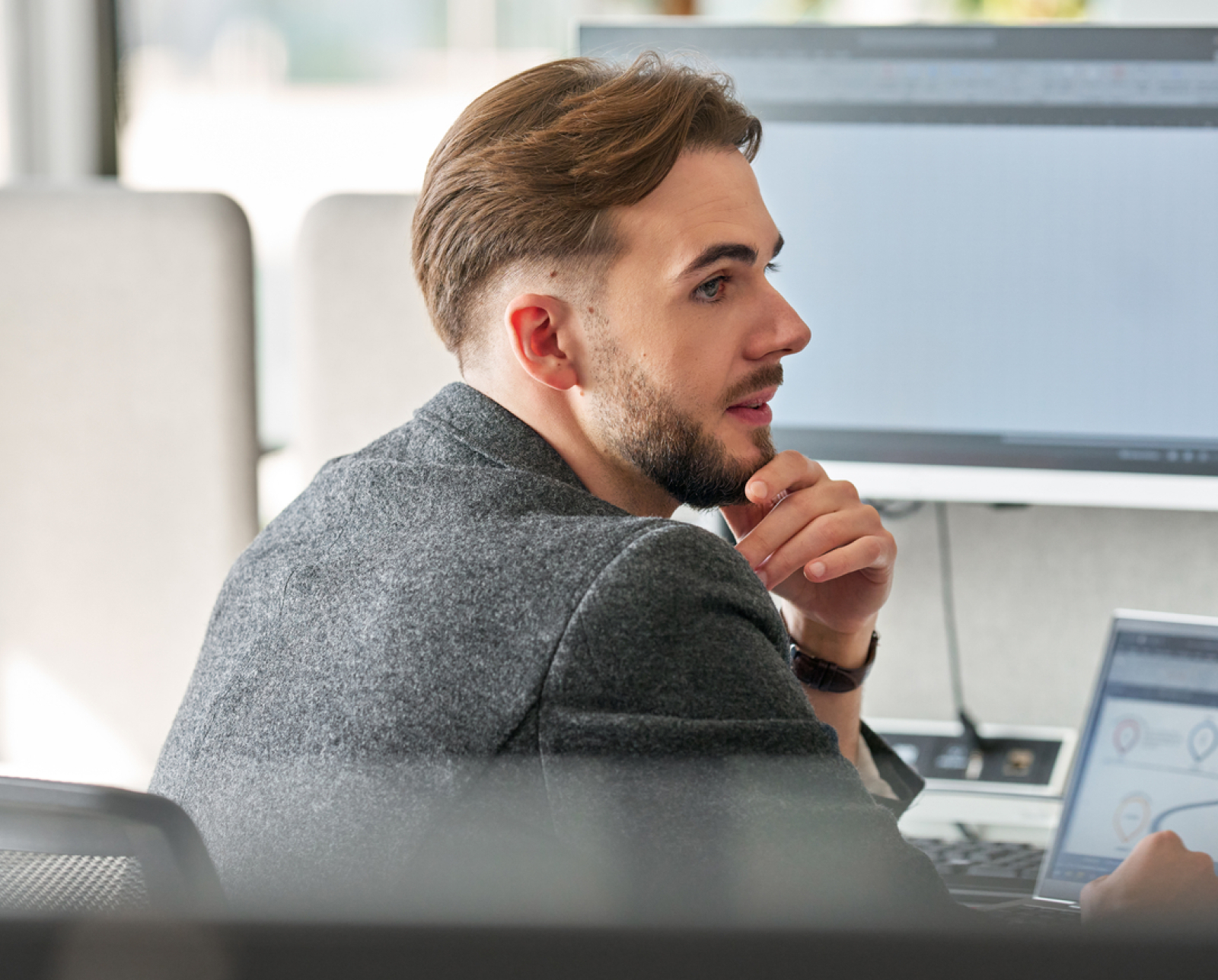 A man with short brown hair and a beard sits at a desk, looking thoughtfully at a computer screen. He rests his chin on his hand and has a laptop open in front of him.