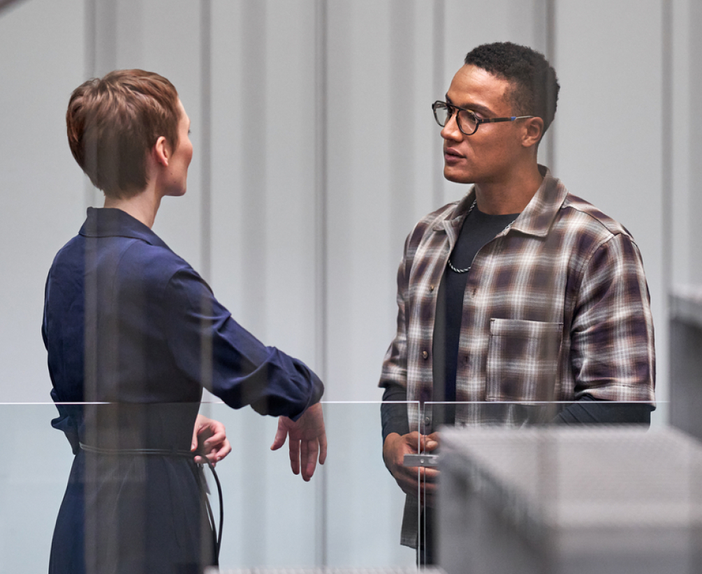 Two people are standing indoors, engaged in conversation. One person wears a plaid shirt and glasses, while the other wears a dark, long-sleeved outfit. They appear to be in a modern, minimalist building.