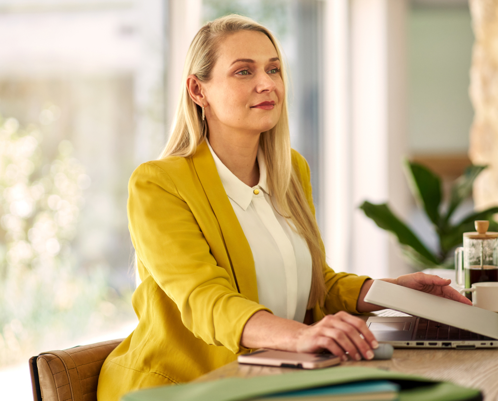A woman with long blonde hair, wearing a yellow blazer and white blouse, sits at a desk using a laptop. She looks thoughtfully into the distance in a bright, modern office setting with plants in the background.
