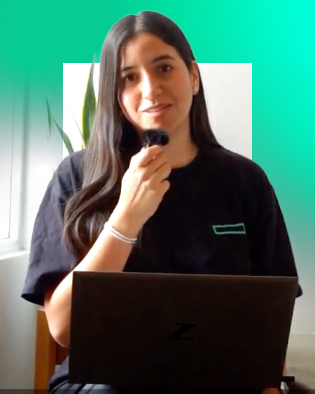 A woman with long dark hair holds a microphone and sits in front of a laptop, wearing a black t-shirt, with a green gradient background and a plant visible beside her.