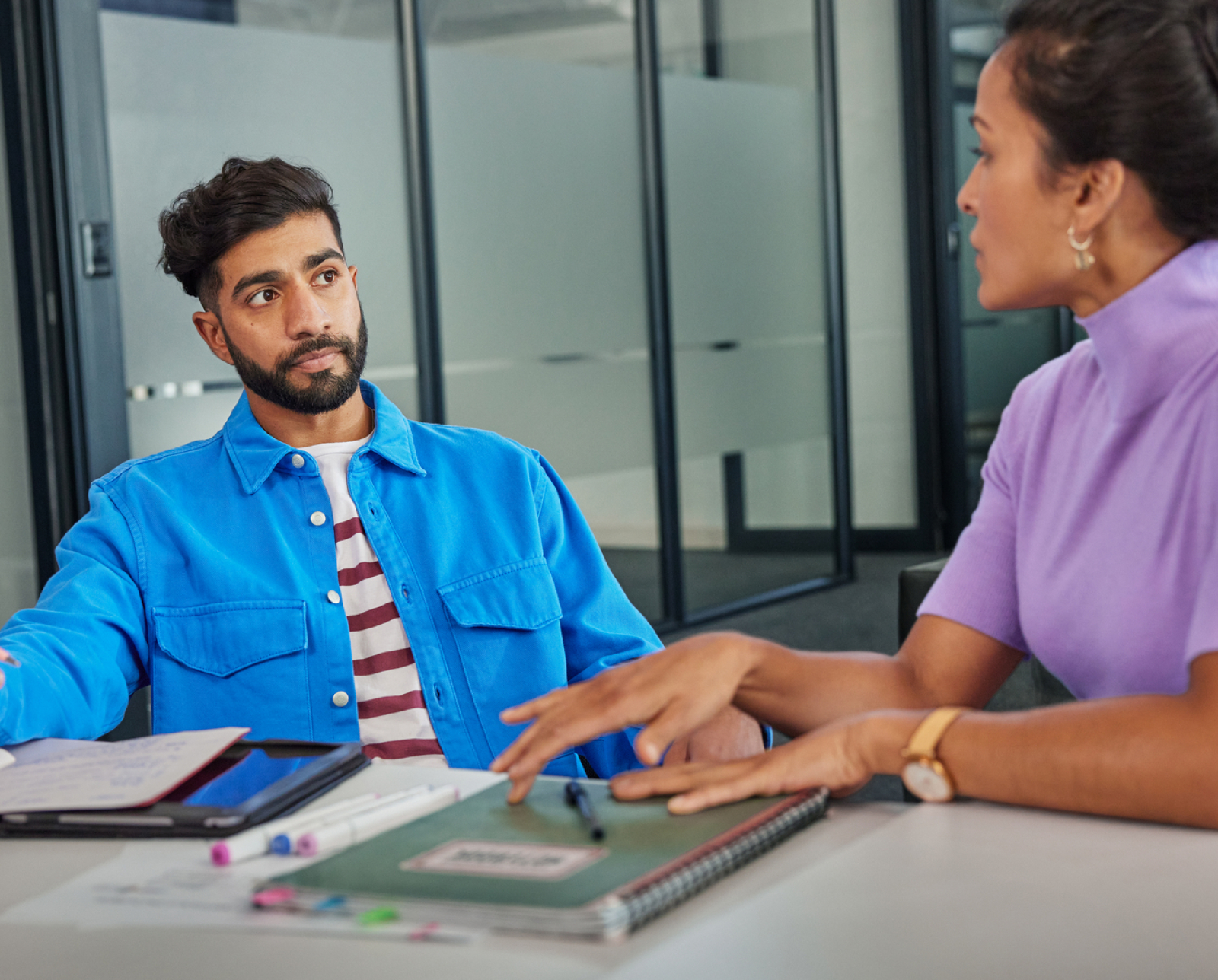 Two people sit at a table with notebooks and pens, engaged in a serious conversation in a modern office setting. The man listens intently while the woman gestures as she speaks.