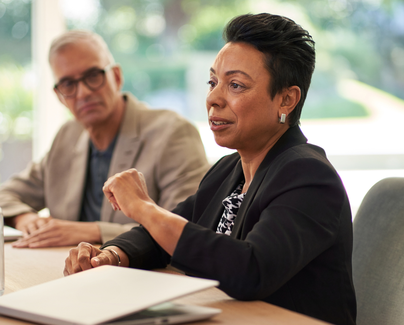 A woman in a black blazer speaks during a meeting, gesturing with her hand, while a man with glasses listens beside her. A laptop and papers are on the table in front of them.