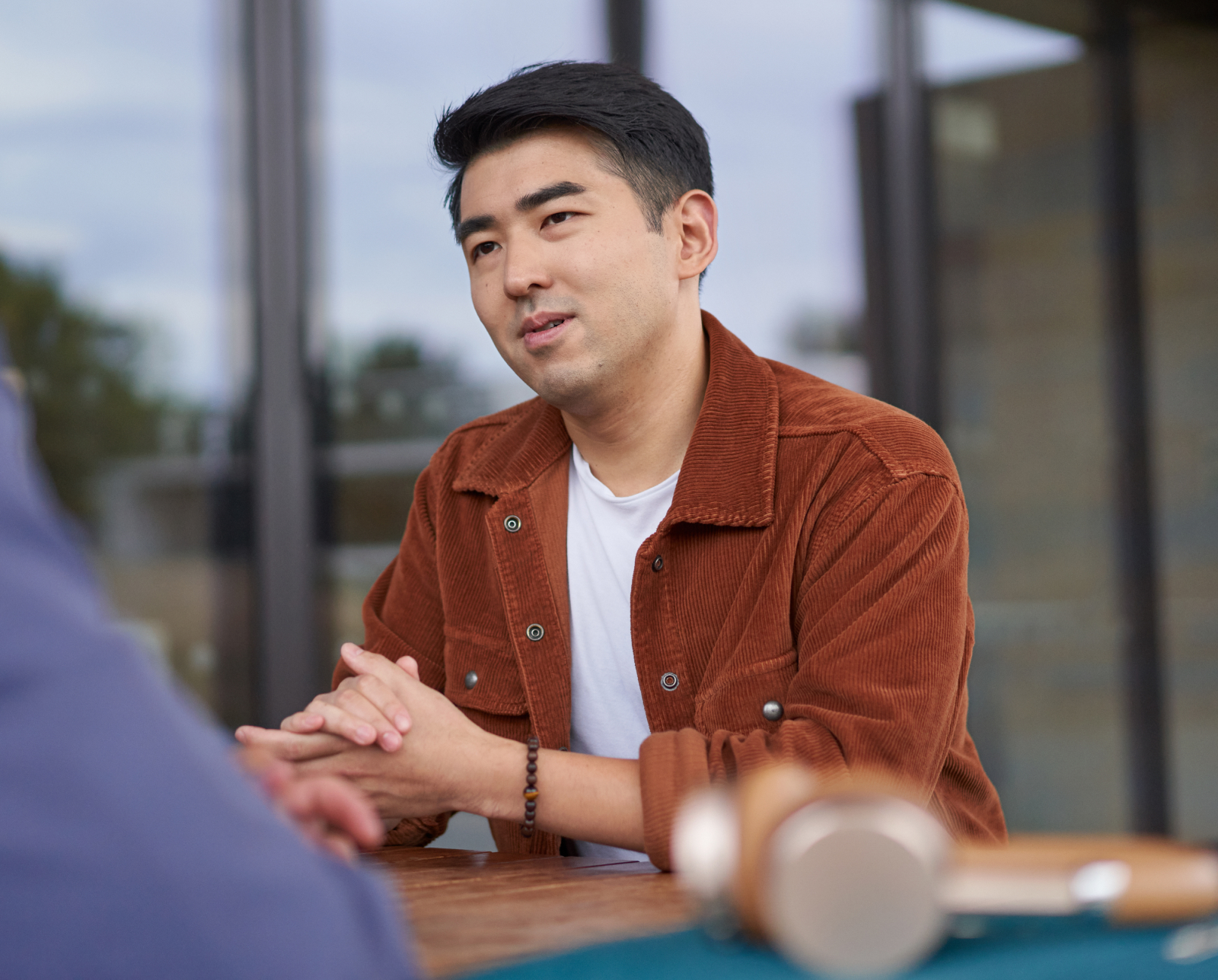 A man in a rust-colored jacket and white shirt sits at a table, hands clasped, listening attentively. A blurred gavel rests on the table in the foreground.