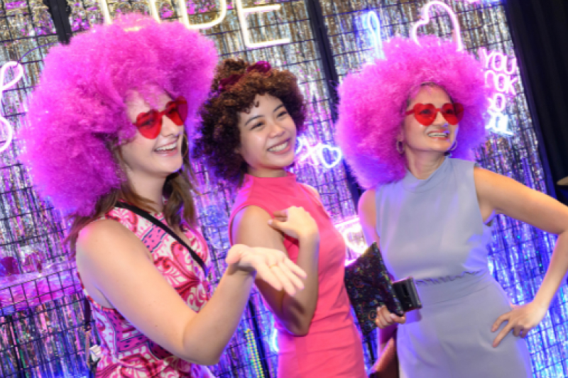 Three people wearing large pink and purple wigs, heart-shaped sunglasses, and colorful outfits pose and smile in front of a neon-lit backdrop. The scene is playful and festive.