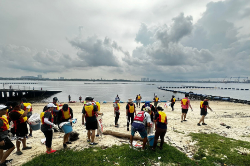 A group of volunteers wearing yellow and red vests clean up litter on a sandy beach by the water under a cloudy sky. Some carry trash bags while others pick up debris and work in teams.