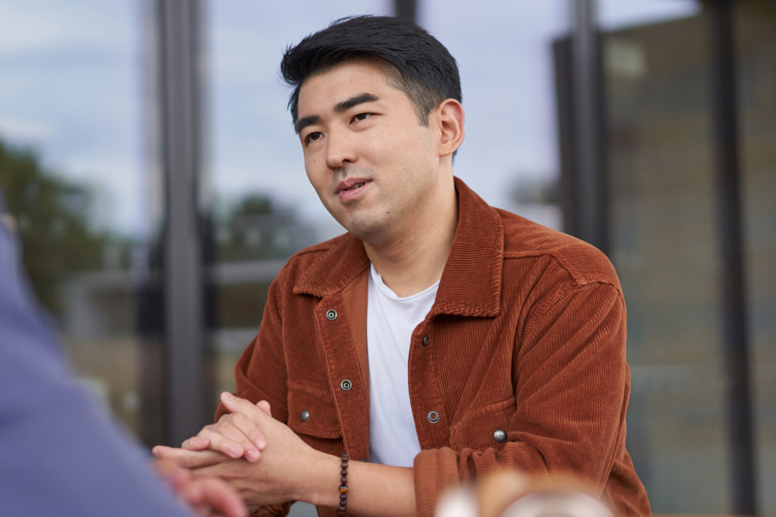 A man in a rust-colored jacket and white shirt sits at a table with his hands clasped, listening attentively during a conversation. Blurred glass windows and outdoor scenery are visible in the background.