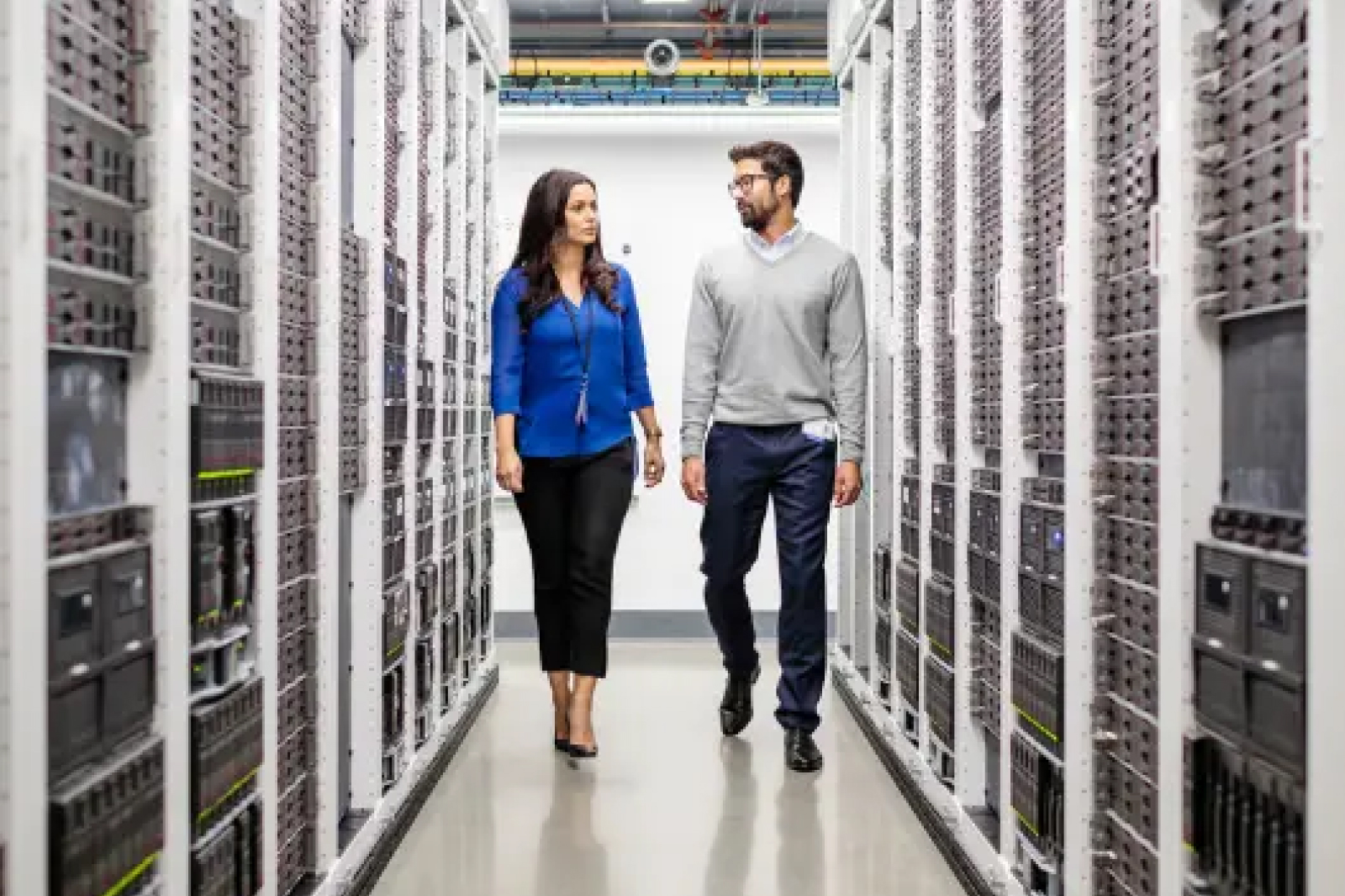 Two people walk through a server room lined with tall racks of computer servers on both sides. They appear to be discussing something as they move down the aisle.