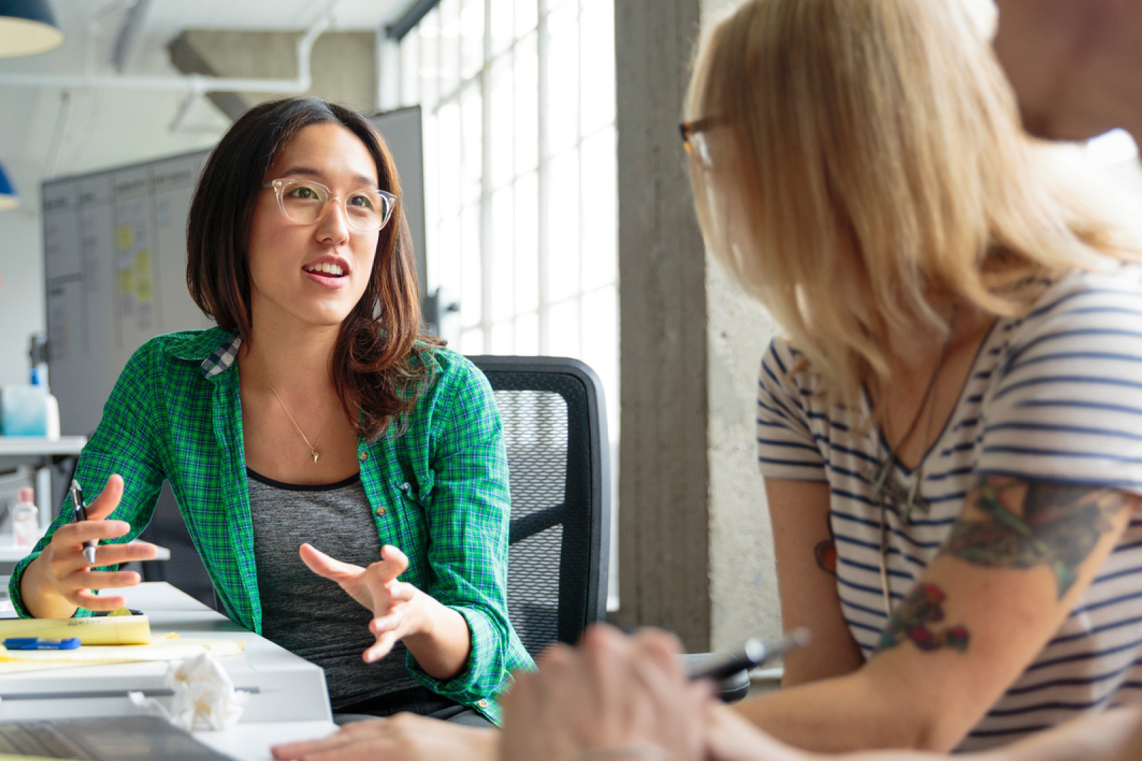 Two women sit at a desk in a bright office, engaged in conversation. One gestures while speaking, and the other listens attentively. Papers and a laptop are on the desk.