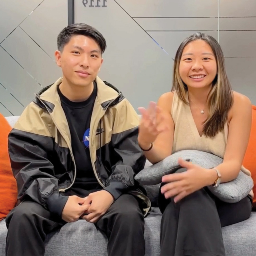 Two young adults sit on a gray couch. The man on the left wears a beige and black jacket and smiles slightly. The woman on the right, in a sleeveless beige top, smiles and gestures with her hand, holding a gray cushion.