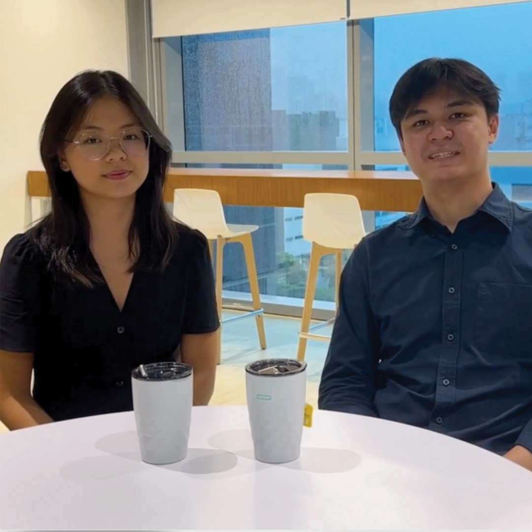 Two people sit at a round white table with two drinks in front of them. They are indoors by large windows, with bar stools and a rainy cityscape visible in the background.