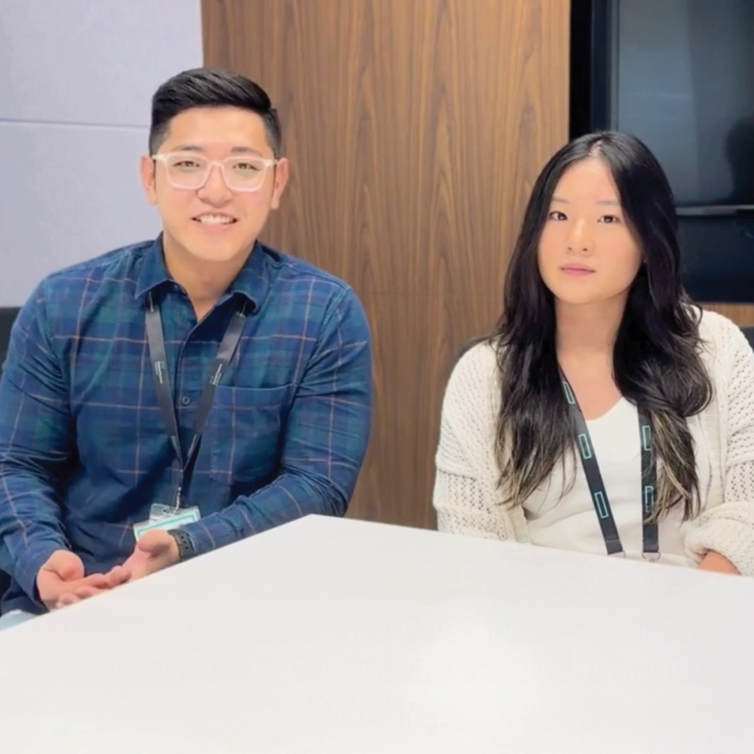 Two people with ID badges sit at a white table in an office setting. The man on the left wears glasses and a blue plaid shirt, while the woman on the right has long dark hair and wears a white cardigan.