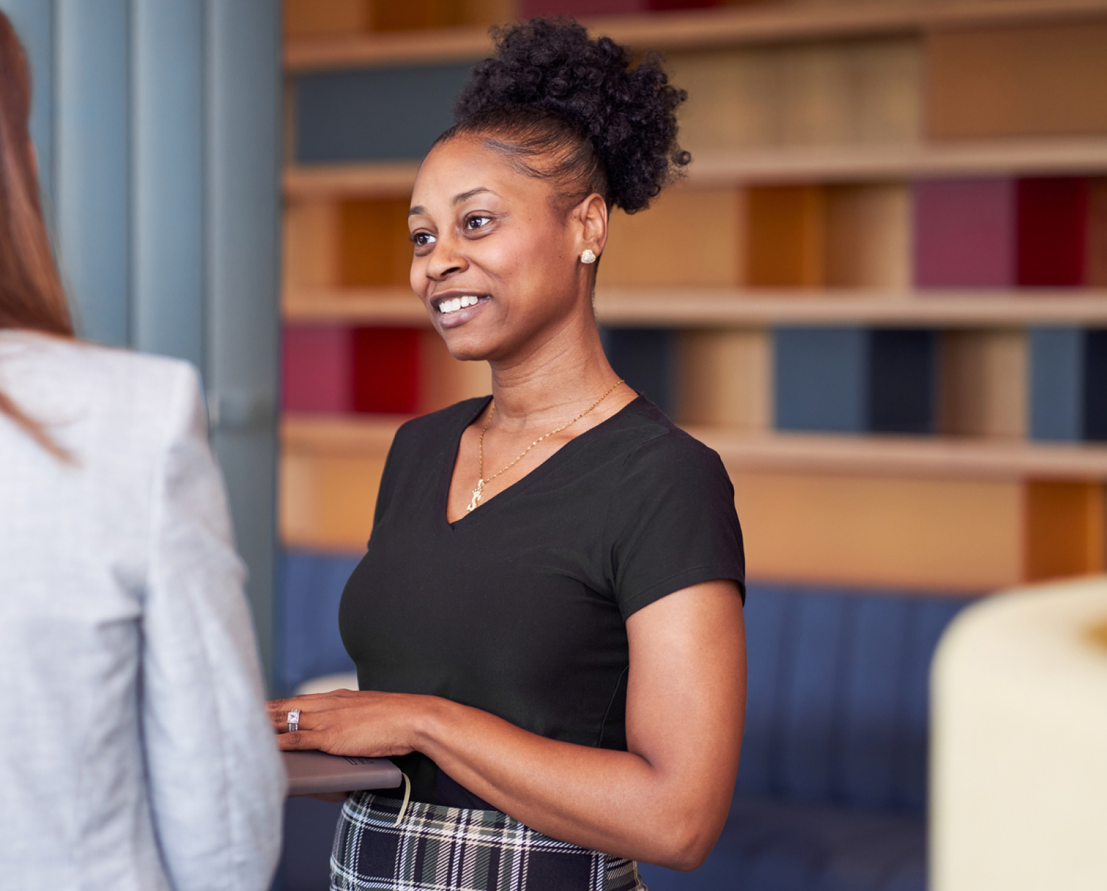 A woman with curly hair in a bun, wearing a black top and plaid skirt, smiles while talking to another person indoors with colorful shelves in the background.